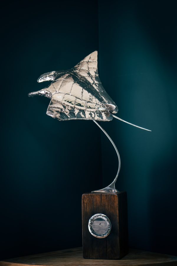 Small manta ray sculpture crafted from metal, displayed on a wooden base.