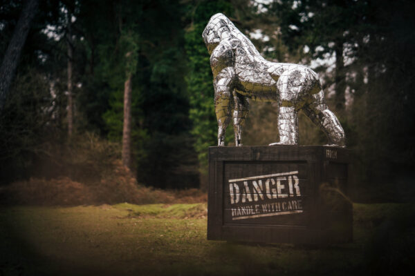 Playful stainless steel gorilla sculpture perched atop a wooden cage labeled "Danger: Handle with Care."